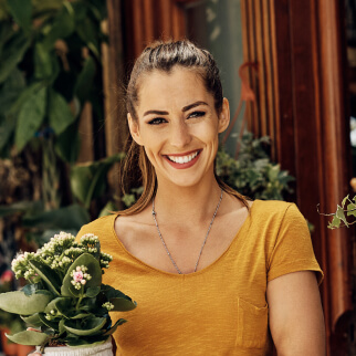 A smiling woman in a yellow shirt holds a potted plant, standing in front of a wooden structure with greenery in the background.