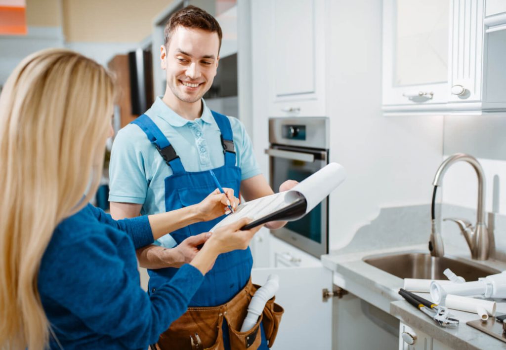 A contractor and a client discuss plans while standing in a modern kitchen. The contractor is smiling and holding a clipboard, while the client points at the document.