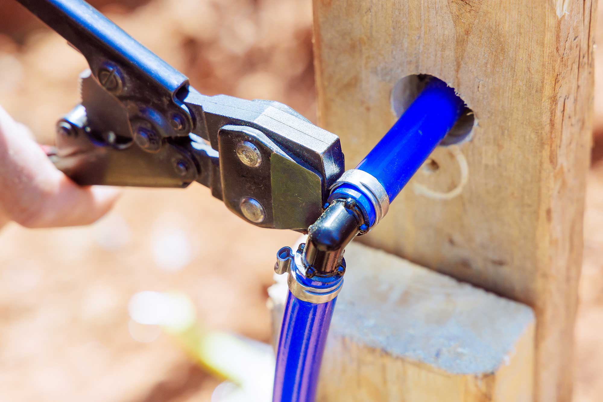 A plumber uses a tool to secure a blue pipe as it connects to a wooden structure. The scene highlights the process of pipe repair to ensure proper water flow.