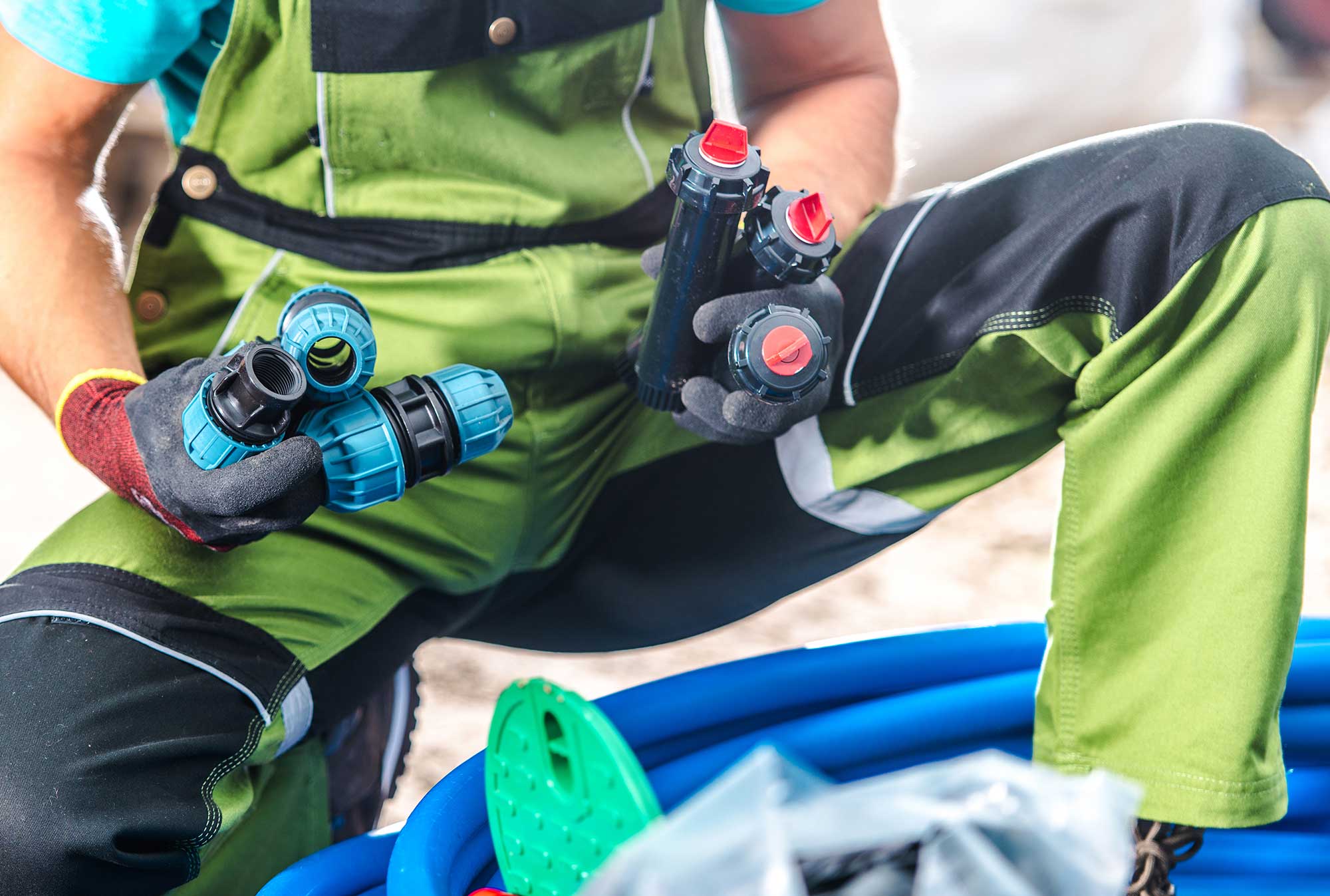 A plumber in green overalls holds two leak detection tools while kneeling beside blue hoses. The focus is on the tools, illustrating the process of identifying plumbing leaks.