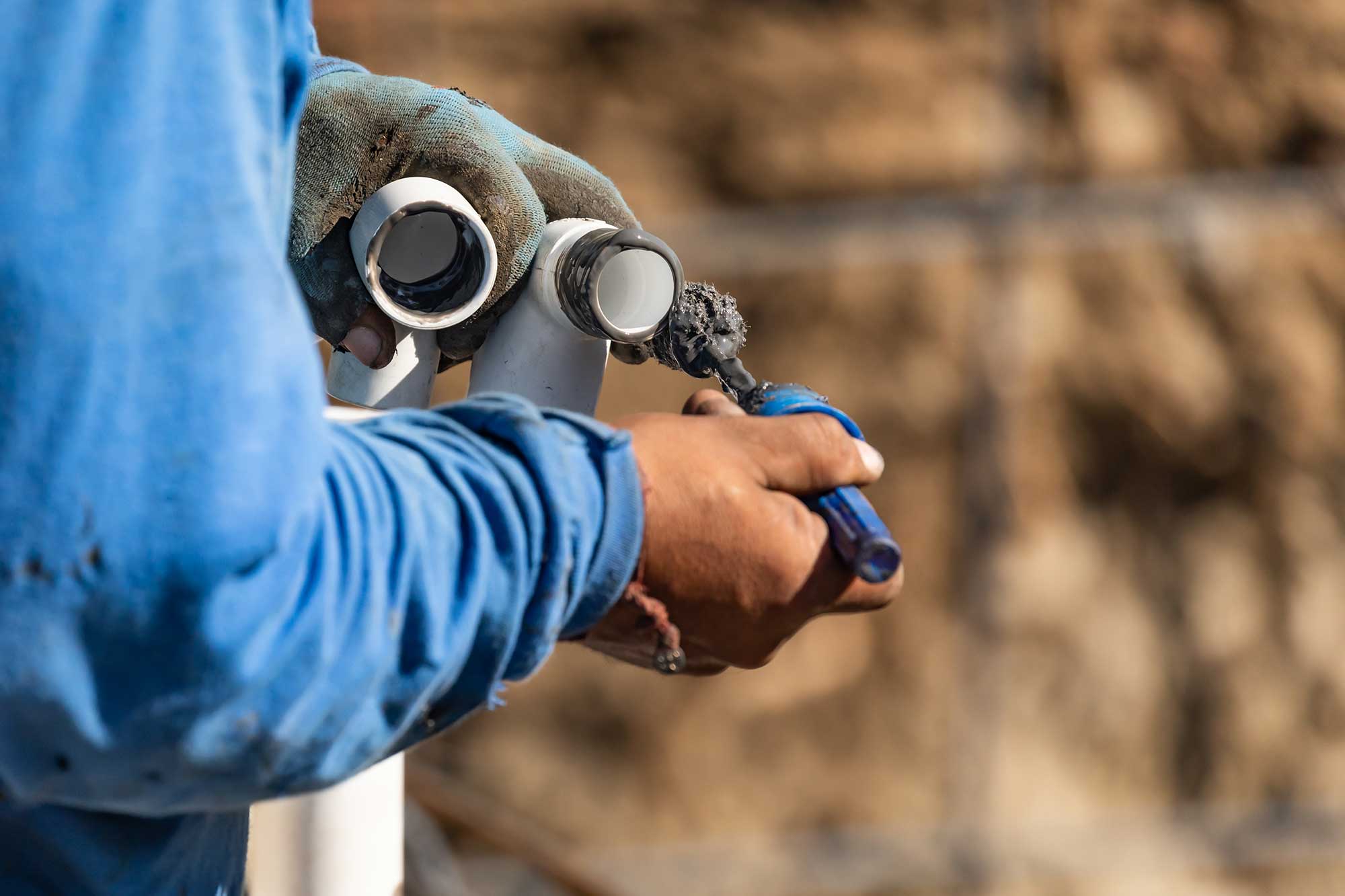 A plumber in a blue shirt inspects plumbing pipes with tools in hand, focusing on leak detection. The background shows a construction site, indicating ongoing work.