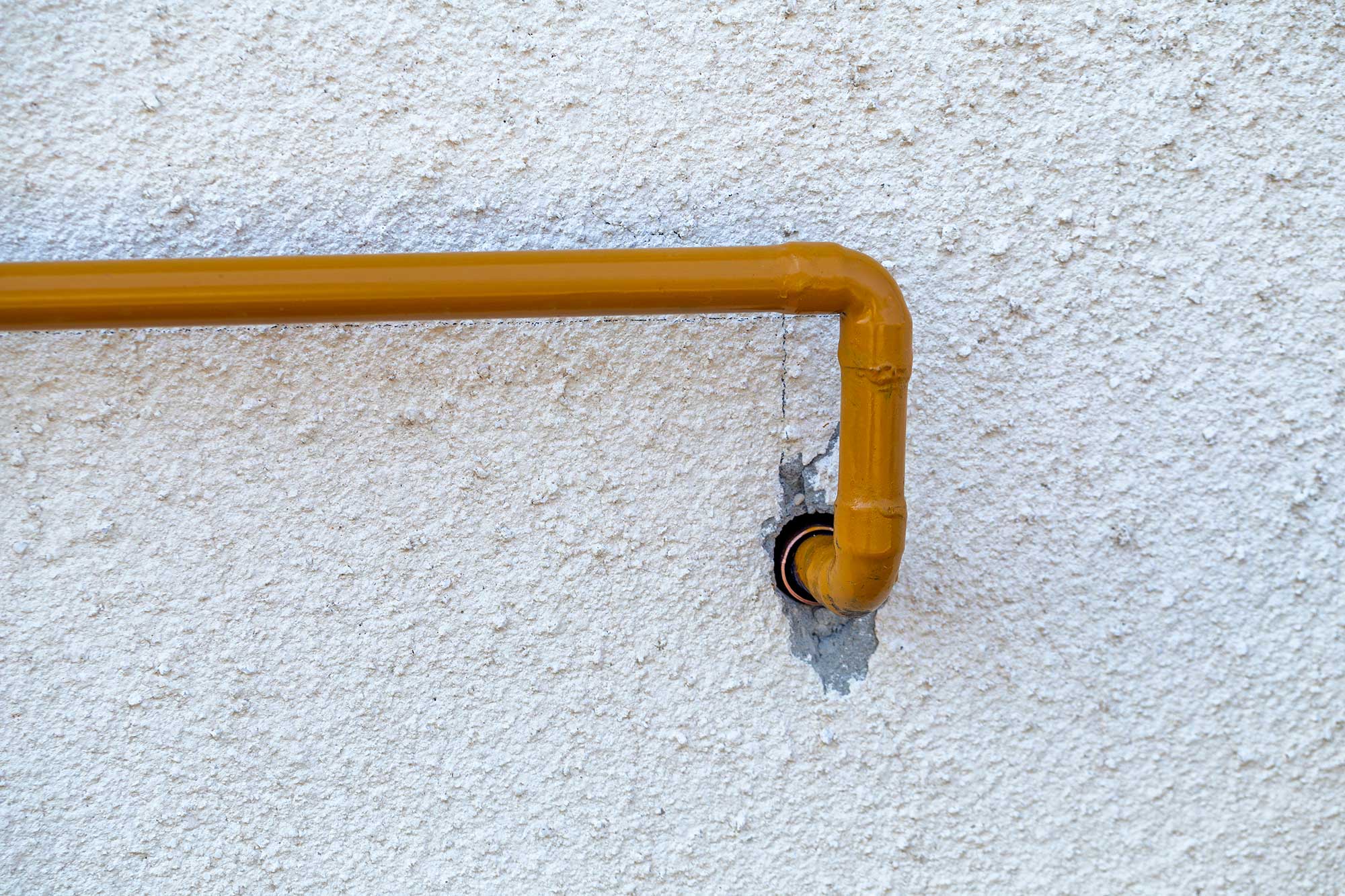 A yellow gas pipe against a textured white wall, bending at a right angle into a hole. The wall has a slightly rough, unfinished surface.